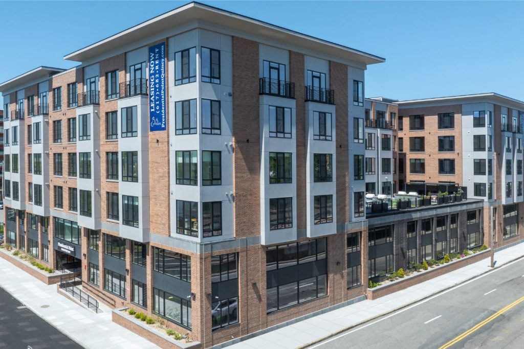 Corner of large condo and mixed use residential project in Quincy Ma, featuring brick and grey facade and first floor parking.